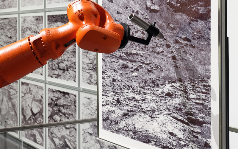 »the native picture« von robotlab. Zu sehen ist ein orangener Roboterarm, der ein schwarz-weiß Bild von einer dürren Landschaft zeichnet.