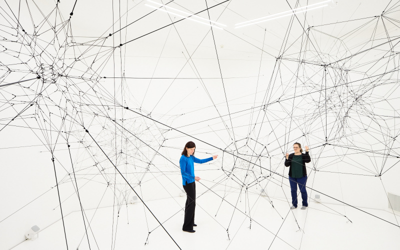 »Algo-r(h)i(y)thms« von Tomás Saraceno. Zu sehen ist ein großes Netz aus Fäden in einem weißen Raum. Zwei Personen stehen in diesem Raum und berühren diese Fäden.