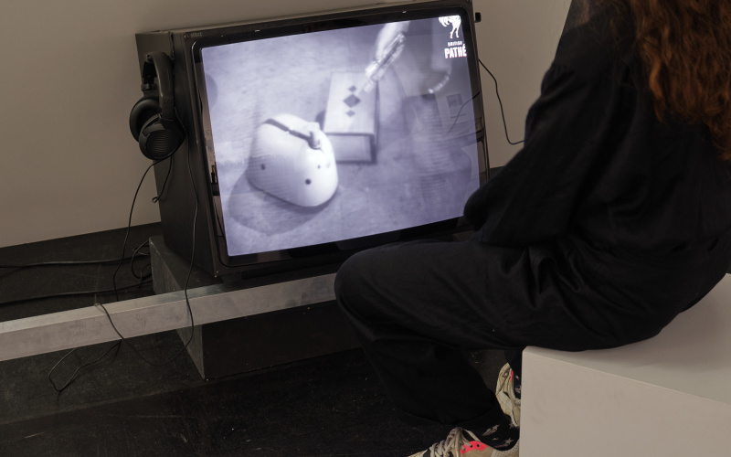 »ELMER – (ELectro-MEchanical Robot)« von William Grey Walter. Zu sehen ist eine Person, die auf einen schwarz-weiß Röhrenfernseher schaut. Sie trägt Kopfhörer und sitzt auf einem weißen Hocker.