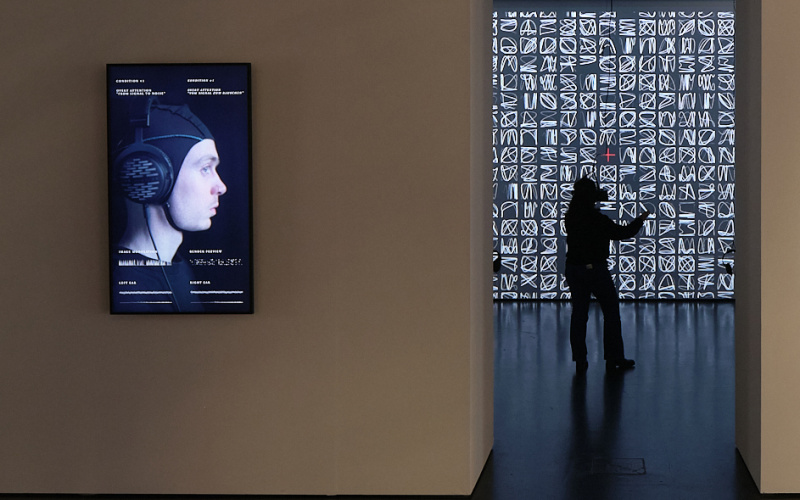 »AUTOPOIESIS« von ATELIER-E. Es sind zwei Räume, verbunden durch einen Durchgang zu sehen. An der Wand des vorderen Raumes befindet sich ein Bildschirm, auf dem eine Person mit einer Haube abgebildet ist. Im zweiten Raum steht eine Person mit VR-Brille.