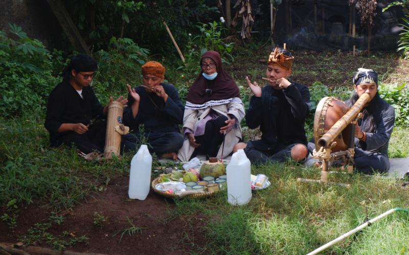 You can see five people sitting on the ground in the forest. In front of them is a tray with cups and coconuts. The people have objects made of wood in their hands and mouths.