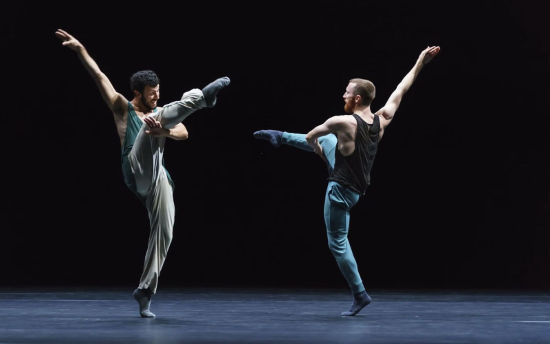 Zwei Tänzer der Choreografie »Friends of Forsythe«. Sie stehen sich gegenüber auf einer schwarzen Bühne und heben kunstvoll ihre Arme und Beine in die Höhe.