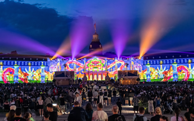 The façade of the baroque palace in Karlsruhe can be seen. The winning entry of the BBBank Award 2023 "BhinnekaExpress", 2023, The Fox, The Folks is projected. The castle glows in bright colours and laser beams shine into the sky. 