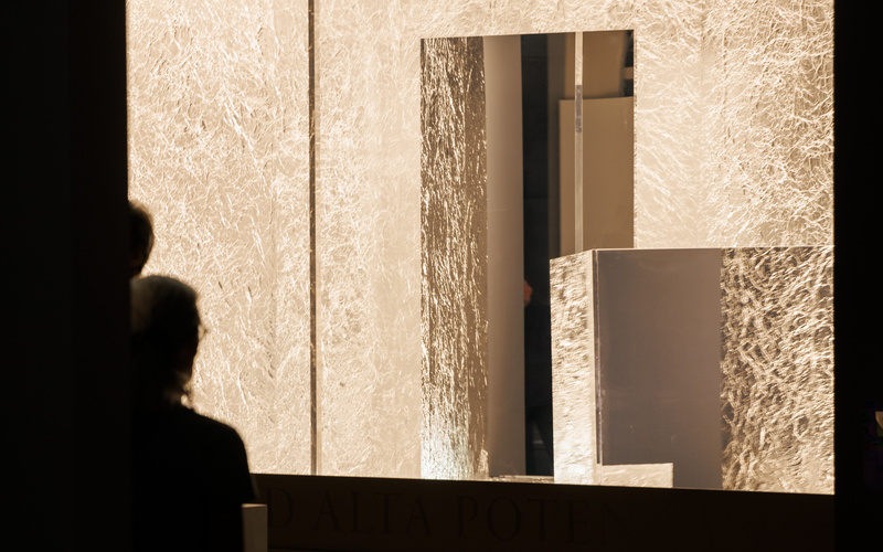 On view is a dark room containing a bright work of art that appears embedded in the wall and is brightly illuminated by lights and mirrors.