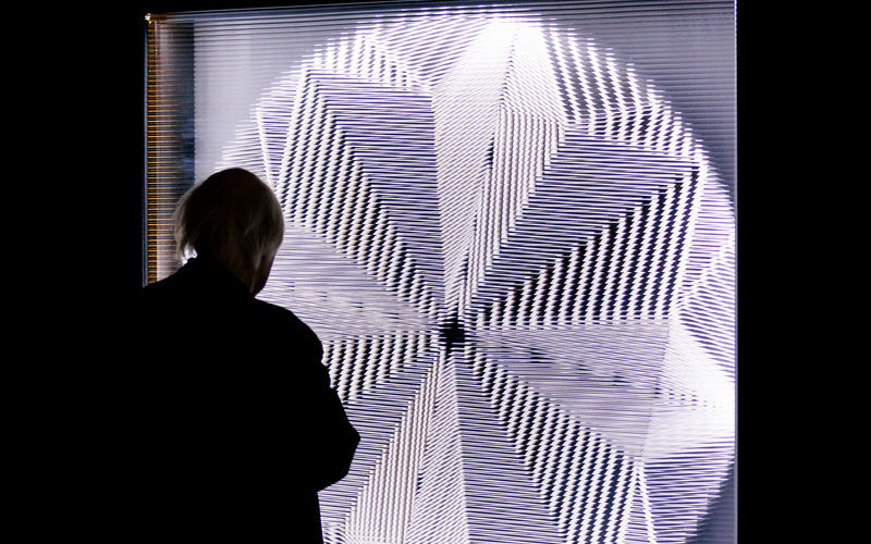 An elderly man stands in front of a round luminous disc, which creates a pattern because the glass in front of it is cut, and looks at it.