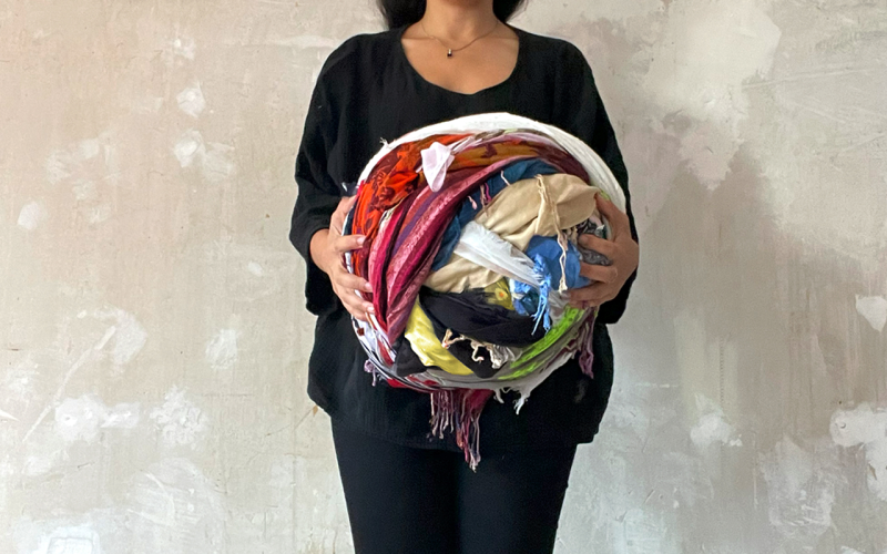 A woman holds colorful scarves in her hands. Next to her, various scarves are connected in a snail shape on the floor.