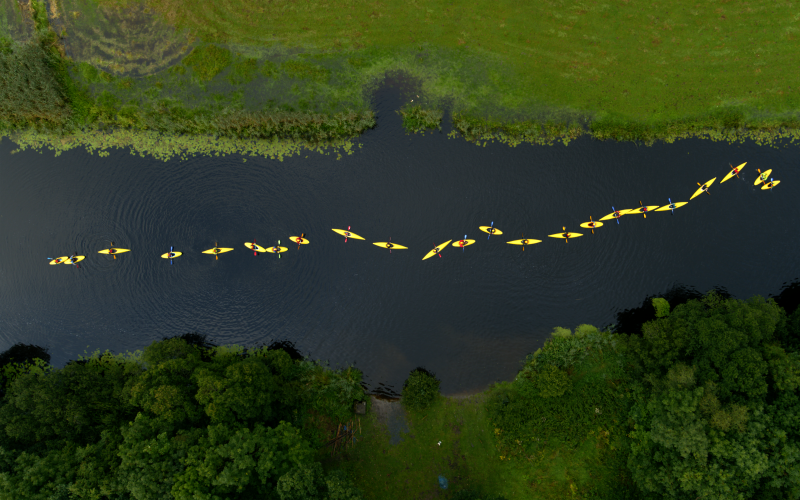 You can see a green landscape with a river meandering through it. The picture shows a bird's eye view of nine people in yellow rain jackets and yellow canoes paddling along the river.