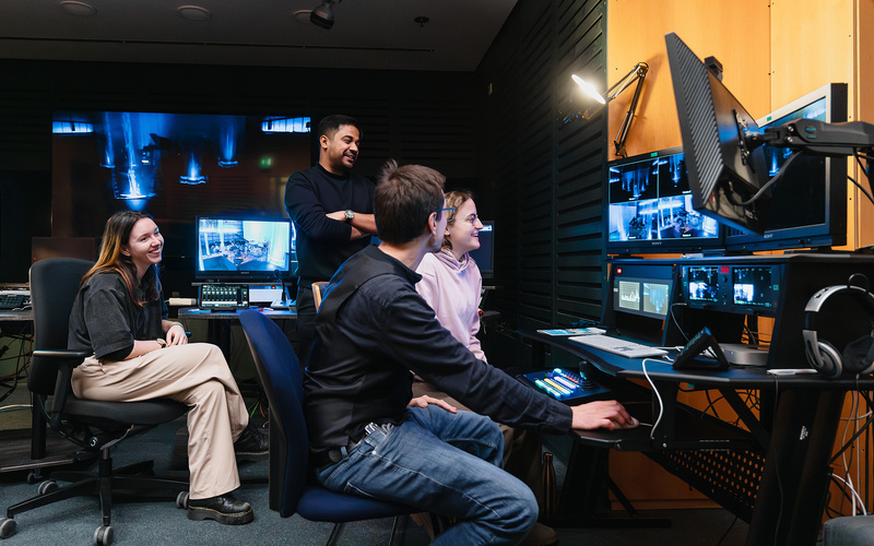 Das Bild zeigt die Mitarbeiter:innen des Videostudios im Regieraum auf Monitore blickend.
