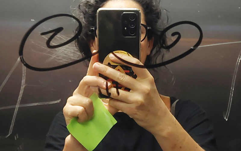 The picture shows a dark-haired woman holding her cell phone in front of her face and taking a selfie in a mirror.