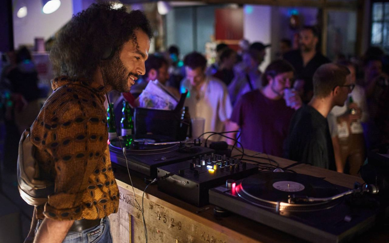 The picture shows a man in half-profile in front of a DJ booth, wearing a colorful shirt and black curls.