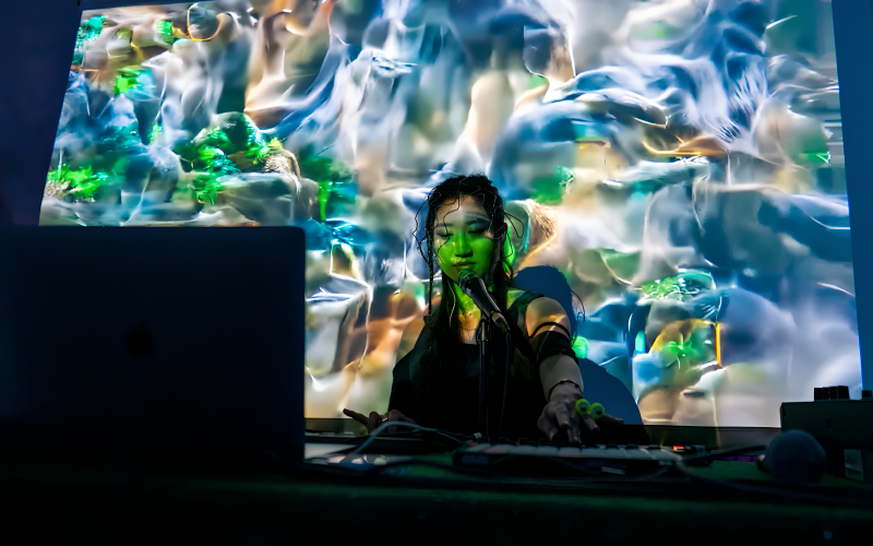 You can see a young woman in front of a DJ booth, she has black hair and a braid and is wrapped in abstract shapes and colors by the lights in the room and a projection behind her.