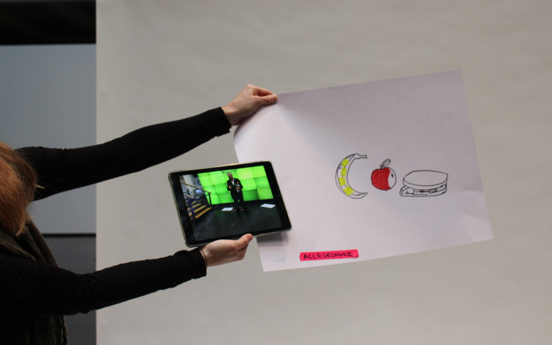 A woman is holding a piece of paper on which a banana, an apple and a sandwich are sketched. On the bottom of the paper the word "Allrounder" written. Also she is holding an iPad with the photo of a man in front of a green screen.