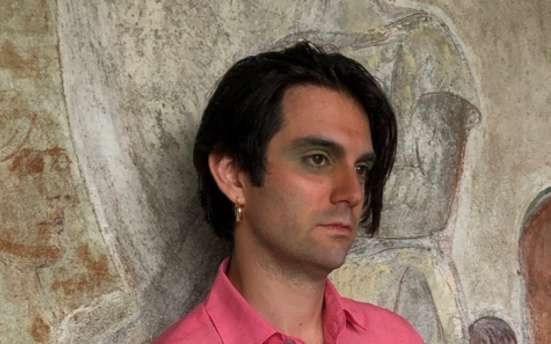 A black-haired man in a pink shirt can be seen leaning against a stone wall and looking out of the picture to the right.