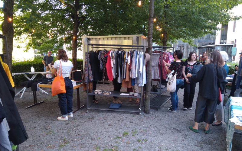 the forecourt in front of the ZKM can be seen, with clothes hanging on many clothes rails and books lying on tables. People are looking at the things.