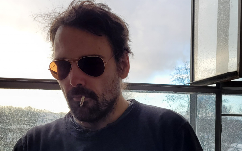 Christian Schlaeffer smoking in front of a window pane wearing a black sweater and sunglasses