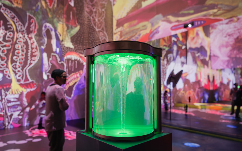 You can see a glass jar that glows green and is filled with water. The water forms a whirlpool. In the background, abstract shapes light up on the walls and a woman walks past.