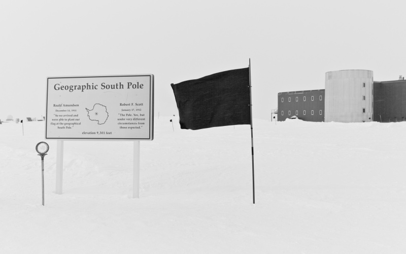 The picture shows a black flag in the snow next to an information board about the South Pole and a house in the background.
