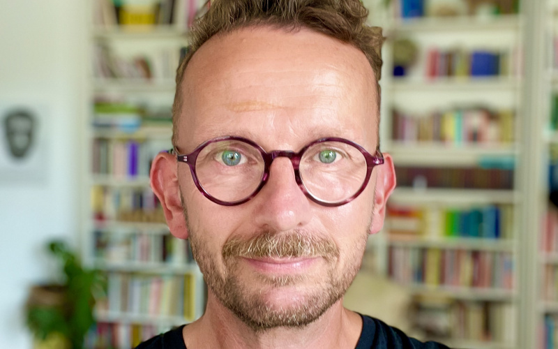 Portrait of a man with round glasses in front of a bookshelf.