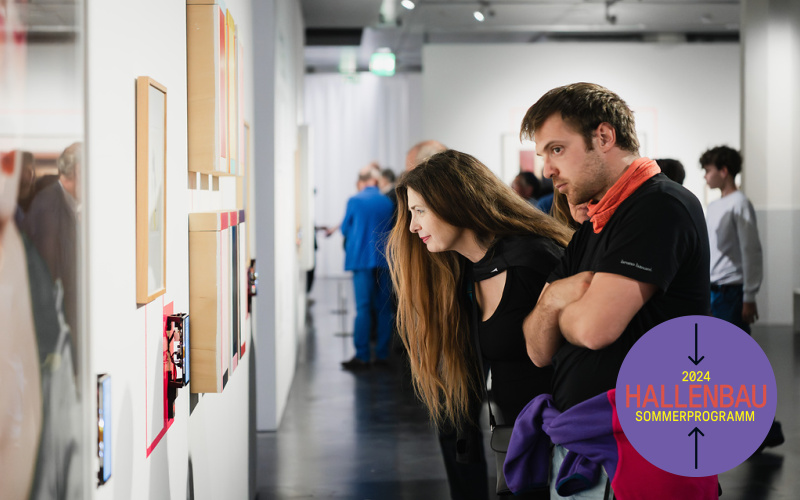 A woman and a man look at pictures on a wall. In the background, several people can be seen walking through the exhibition.
