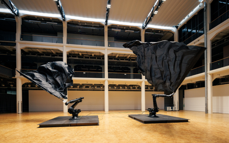 Two robotic arms each wave a large black flag in a spacious atrium