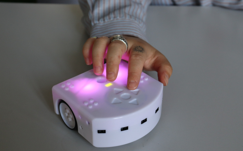Eine Hand bedient einen Thymio-Roboter, der auf einem Tisch liegt.