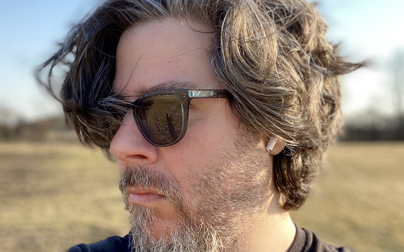 Das Bild zeigt einen Mann mit dunklen Haaren und grauem Bar von der Seite. Eine Sonnenbrille verdeckt seine Augen. Im Hintergrund ist ein Feld und blauer Himmel.