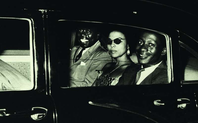 An old black and white photo from the 1950s, two men and a woman sitting in a black car.
