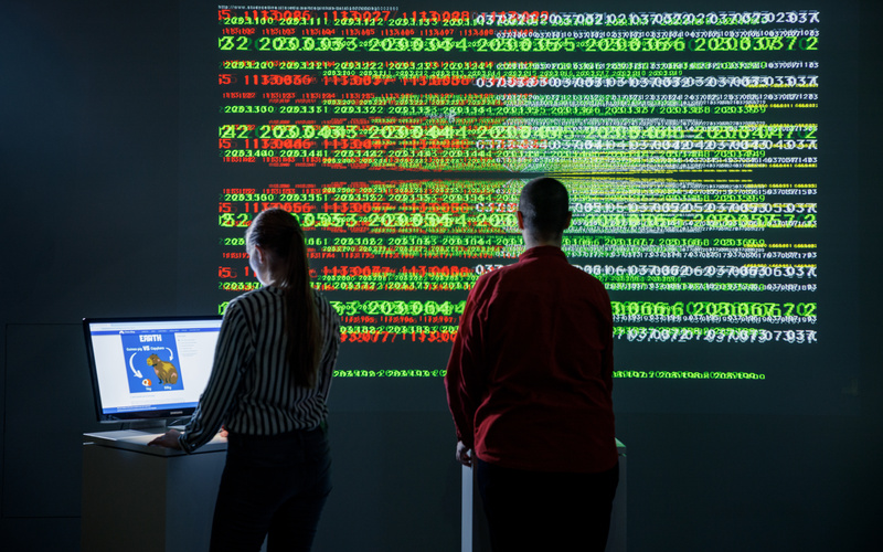 Eine Person steht in einem dunklen Raum vor einer Wand voller grüner und roter Ziffern. Links davon steht eine andere Person an einem Computerbildschirm.