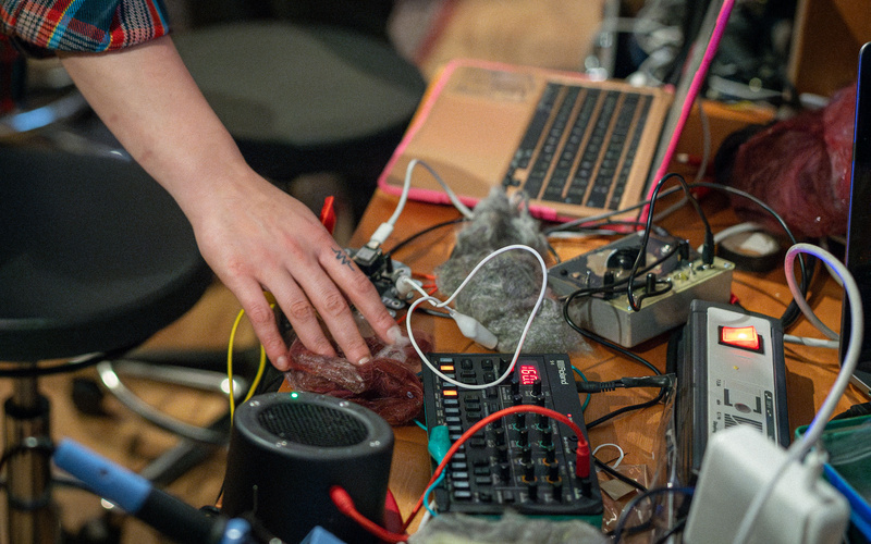 The image shows a hand operating various electronic musical instruments and synthesizers on a table with cables, a laptop, and other audio equipment.