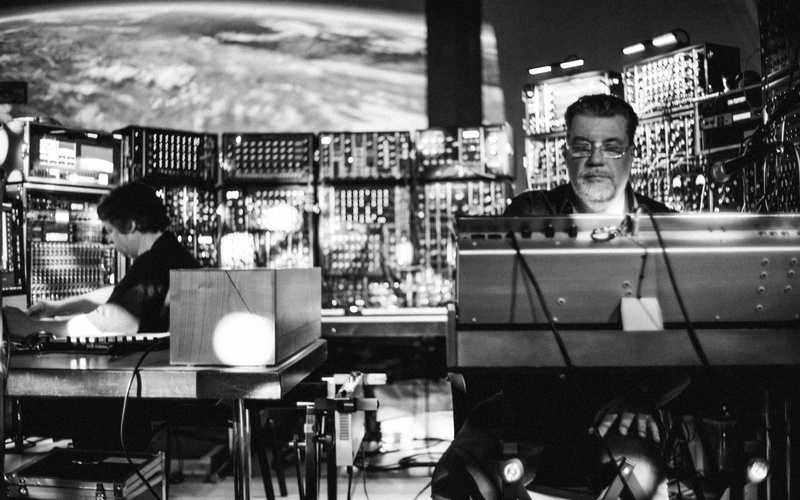 Das Bild zeigt zwei Personen, die von modularen Synthesizern umgeben sind.