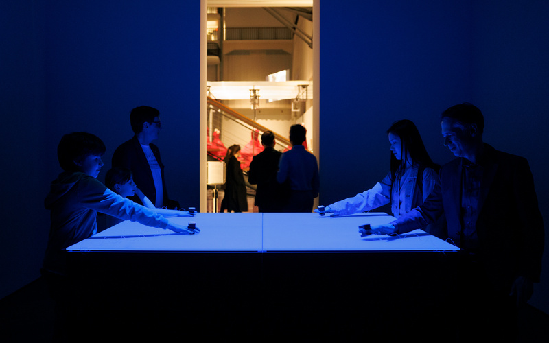 People hold their hands above a white tabletop in a room lit in blue.