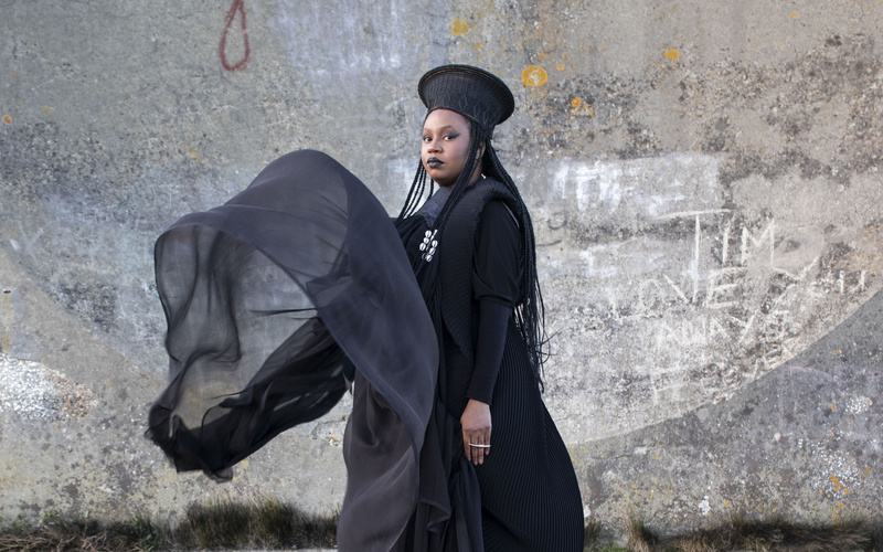 A woman in a black flowing dress stands in front of a concrete wall.