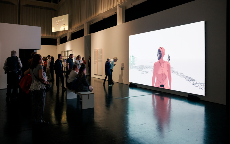 Mehrere Menschen schauen sich in einem Ausstellungsraum eine Videoinstallation an, welche au einem Monitor an der Wand zu sehen ist. 