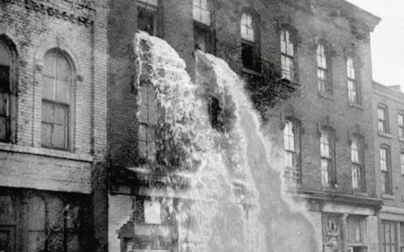 Man sieht ein Fabrikgebäude, aus dessen Fenster ein Wasserfall hervorbricht in Schwarz-Weiß.