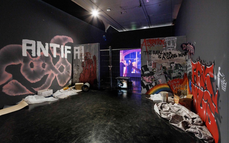 Installation in einem Ausstellungsraum mit politischen Graffiti, Absperrgitter, Projektionsbildschirm und verstreuten Materialien auf dem Boden.
