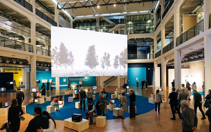 Visitors view a multimedia exhibition featuring vintage TVs and a large-scale video projection in an open museum space.