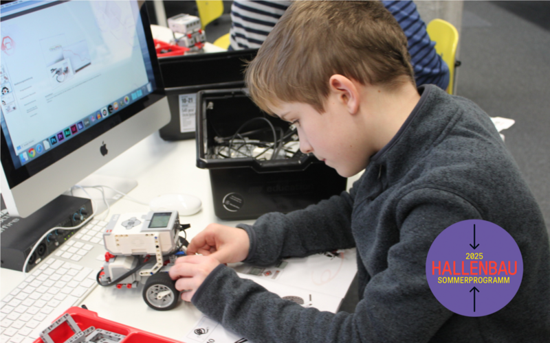 Ein Junge arbeitet an einem Lego-Roboter, den er vor sich auf dem Tisch stehen hat. Um ihn herum stehen Kisten, in denen Einzelteile liegen.