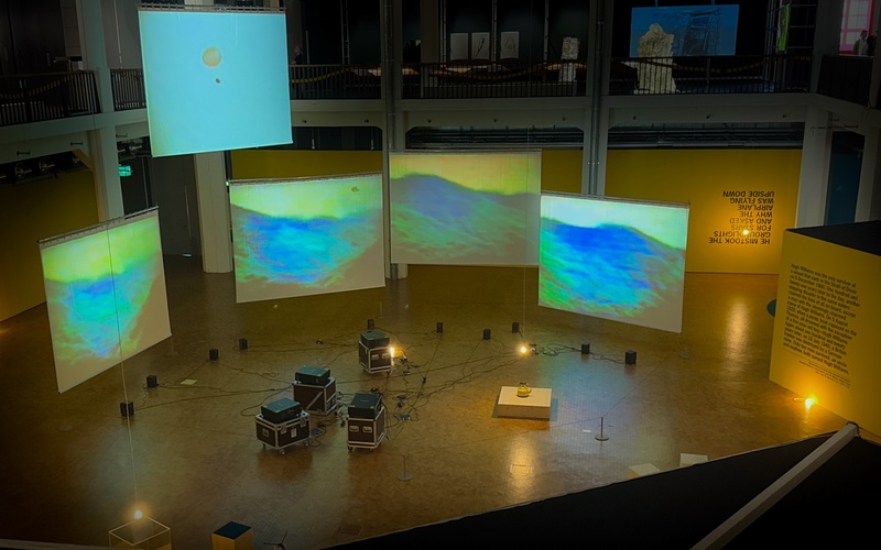 Ausstellungsansicht mit gelben Wänden und beleuchteten Leinwänden.