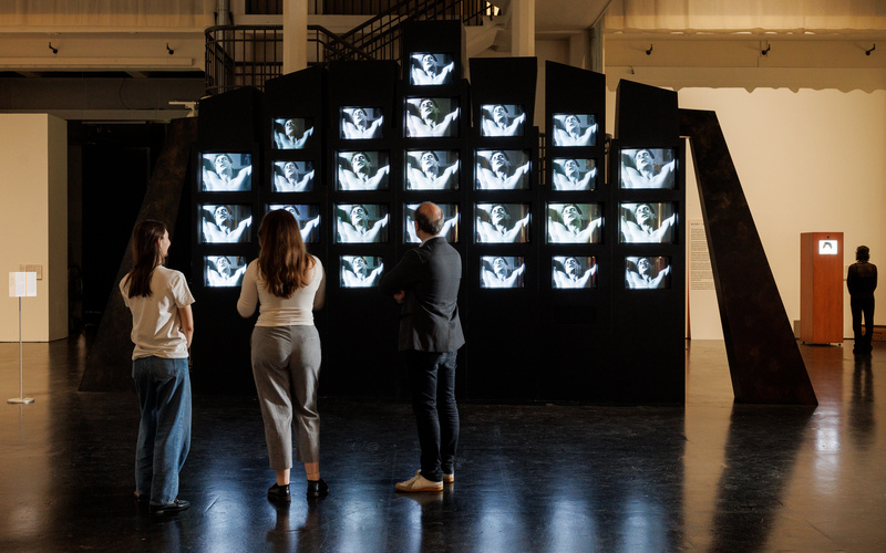 Menschen sind zu sehen, welche vor einer bildfüllenden monumentalen Videoinstallation stehen, welche aus mehrere kleine Bildschirmen, eingerahmt in schwarzem Holz, besteht.