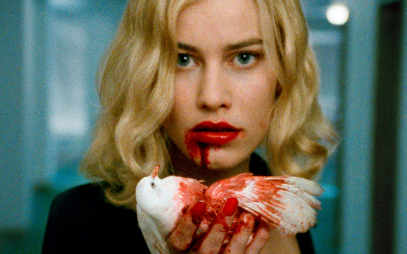 Zu sehen ist eine blonde Frau mit roten Lippen, die einen blutigen weißen Vogel in der Hand hält.  