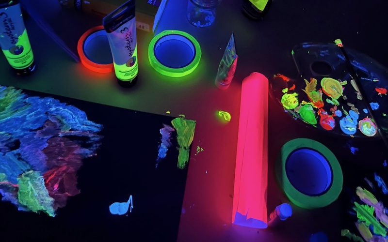 Zu sehen ist ein Tisch im Dunkeln auf dem neonfarbene Bilder und Farbtuben liegen.