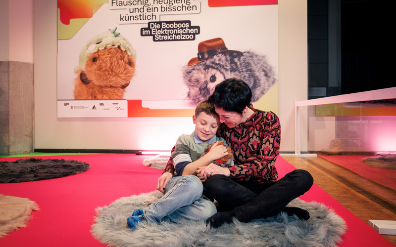 A mother cuddles with her child on the floor, who is holding a robotic guinea pig.