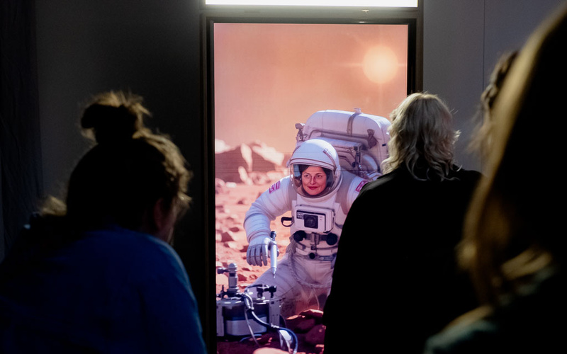 Zu sehen sind mehrere Personen, die vor einem Bildschirm stehen, der eine Astronautin zeigt.