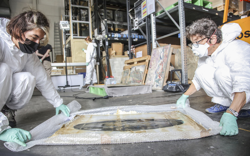 Zu sehen sind zwei Personen, die Masken, Handschuhe und einen Schutzanzug tragen.
