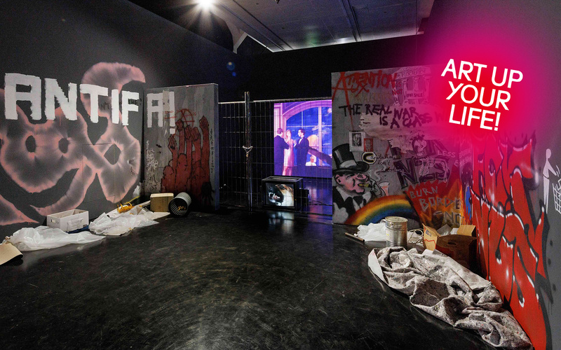 Installation in einem Ausstellungsraum mit politischen Graffiti, Absperrgitter, Projektionsbildschirm und verstreuten Materialien auf dem Boden.
