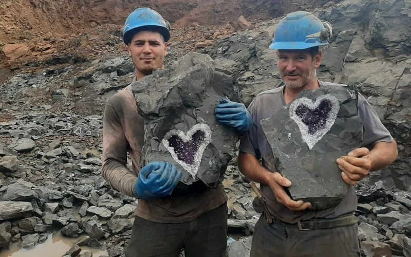 Zwei Personen mit Helmen sind zu sehen, die Gesteinsbrocken in die Kamera halten. 