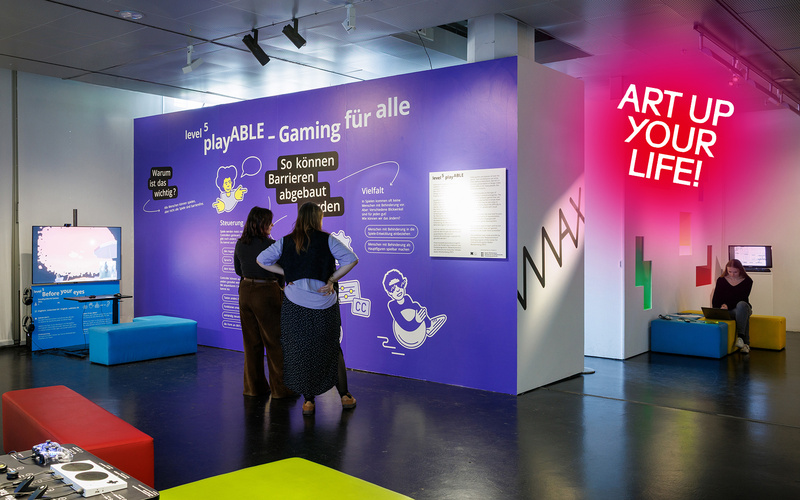 Two people are reading text on a purple wall, one person is sitting on the right in the background.