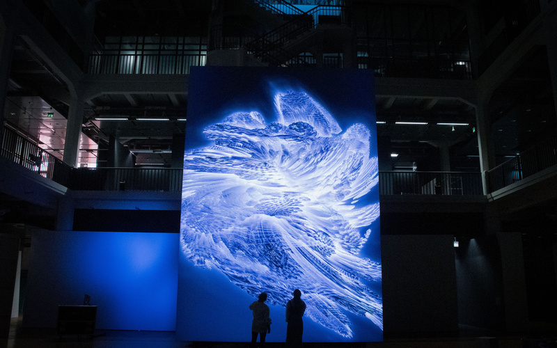 Zwei Silhouetten stehen vor einem riesigen Bildschirm, auf dem ein abstraktes Kunstwerk zu sehen ist.