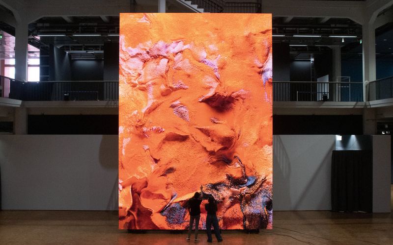 Zwei Silhouetten stehen vor einem riesigen Bildschirm, auf dem ein abstraktes Kunstwerk zu sehen ist.