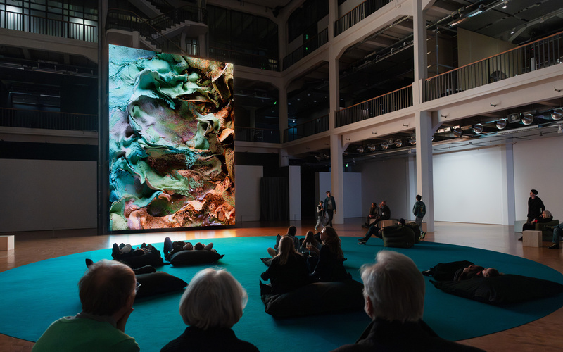 Einige Personen sitzen vor einem riesigen Bildschirm, auf dem ein abstraktes Kunstwerk zu sehen ist.